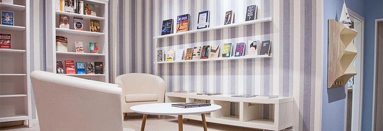 Sitzecke im hinteren Teil des Buchladens bestehend aus zwei weißen Sesseln mit kleinem, rundem Couchtisch. Im Hintergrund und rechts der Sitzgruppe ist eine blau-weiß-gestreifte Wand  mit zwei untereinander angebrachten Regalleisten, auf denen mehrere Büc